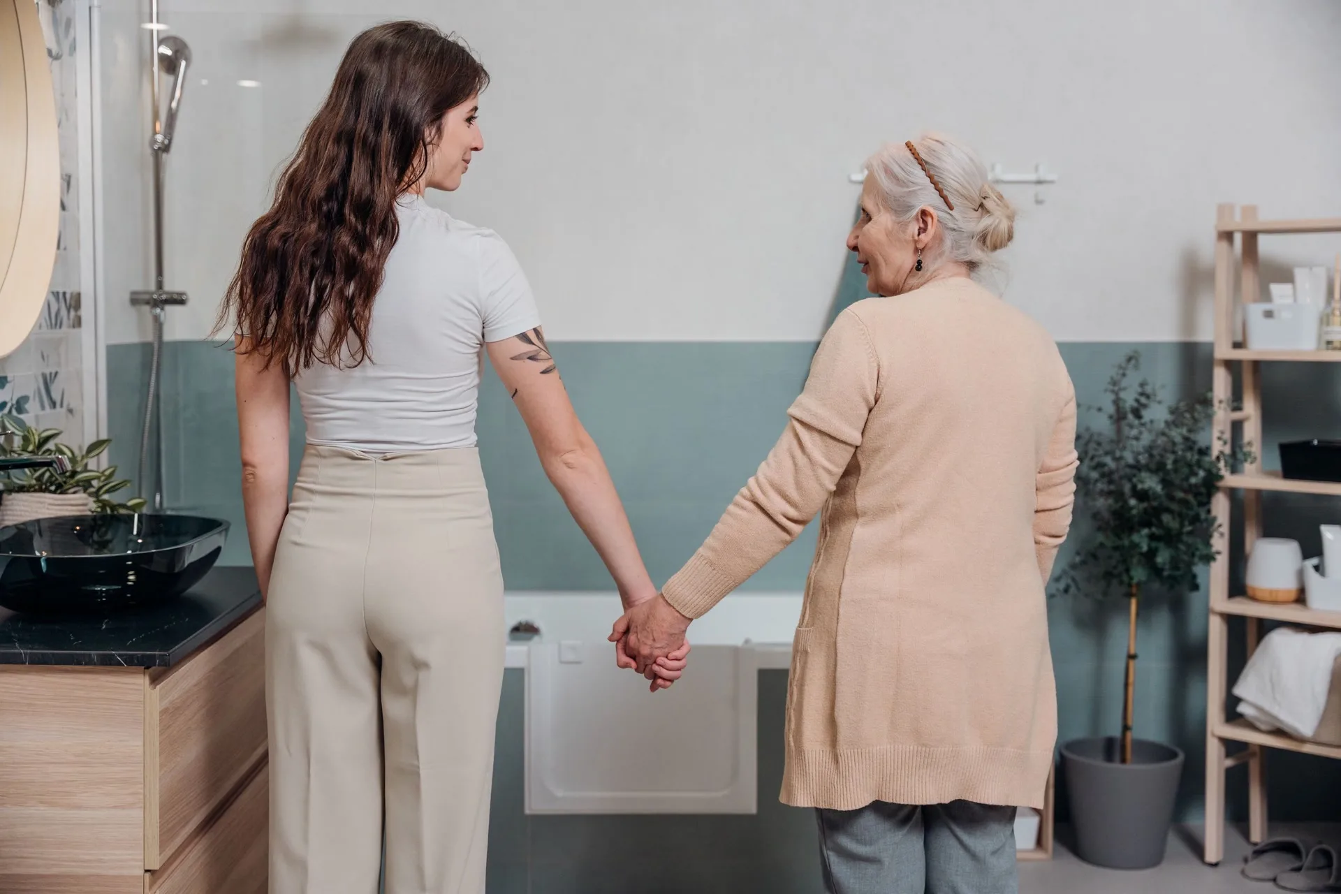 A daughter and her mother stand hand in hand in a bathroom, the bath fitted with a UDOOR® bathtub door behind them